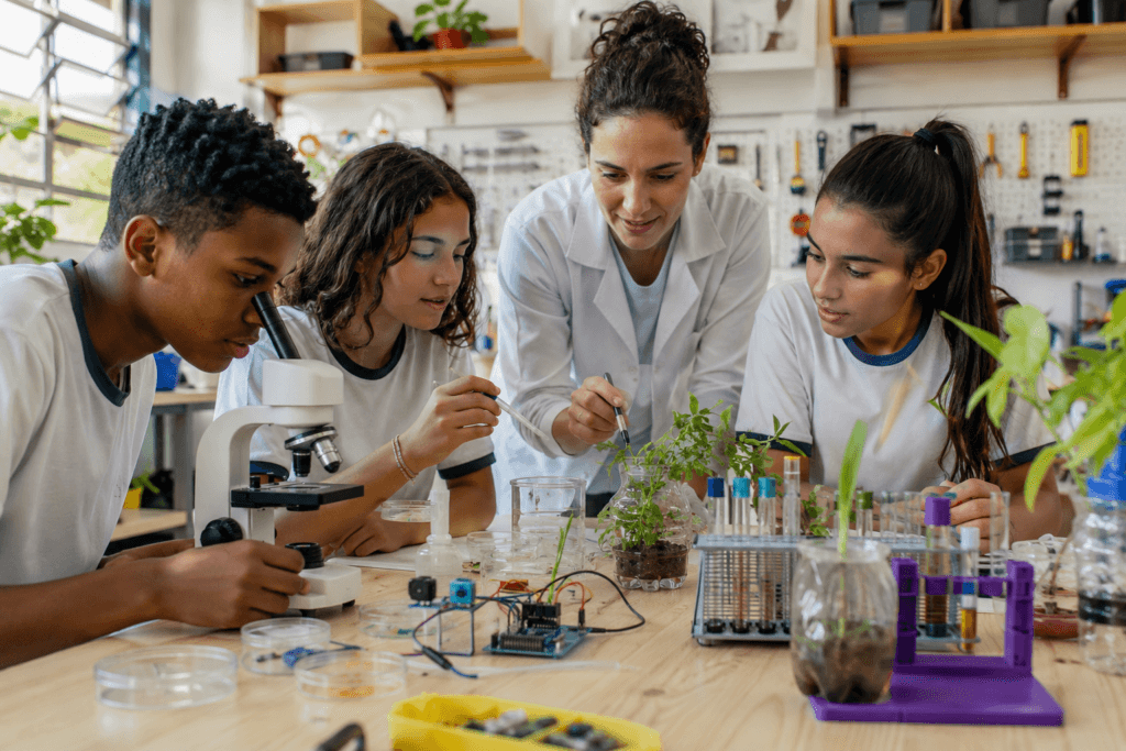 Professor orientando alunos em laboratório de biologia DIY escolar, com microscópio, plantas, tubos de ensaio e componentes maker sobre a bancada.