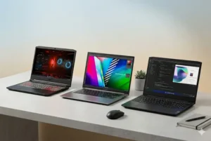 Três laptops modernos estão alinhados em uma superfície de mesa texturizada de concreto, cada um aberto para revelar uma interface de usuário diferente, todos sob iluminação suave. À esquerda, o primeiro laptop tem um teclado com retroiluminação vermelha brilhante e uma interface de sistema operacional escura e angular com elementos gráficos de ficção científica vermelhos. No centro, o segundo laptop apresenta um acabamento de metal prateado e uma tela com um gráfico de usuário criativo colorido e vibrante. À direita, o terceiro laptop tem uma estética preta e exibe código de programação em modo escuro. Um mouse preto sem fio e um caderno cinza com uma caneta estão organizados ordenadamente na mesa, enfatizando um ambiente de trabalho moderno e produtivo. A parede de fundo tem um gradiente suave e a luz natural vem do lado, criando uma sensação profissional e aspiracional.