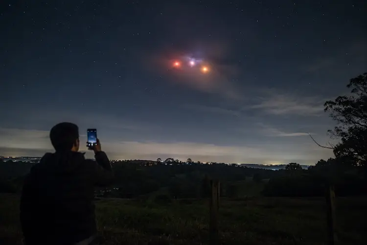 Pessoa grava com celular luzes coloridas misteriosas no c&eacute;u noturno de uma &aacute;rea rural, em cena relacionada ao suposto OVNI no Paran&aacute;.